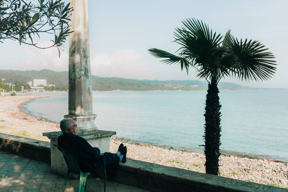 Old man sitting on a chair with his feet on a little wall, watching the sea and a palm tree.